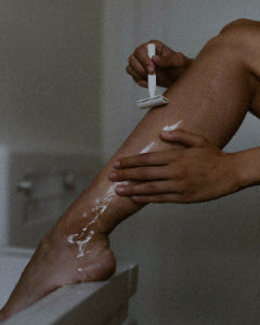 A close-up lifestyle shot of a person shaving their lower leg with white shaving cream, using a white manual razor in a dimly lit bathroom setting.