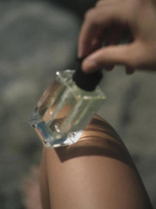 A hand holding a clear glass perfume bottle with a black dropper cap, applying a clear liquid to a person's leg in a softly blurred outdoor setting.